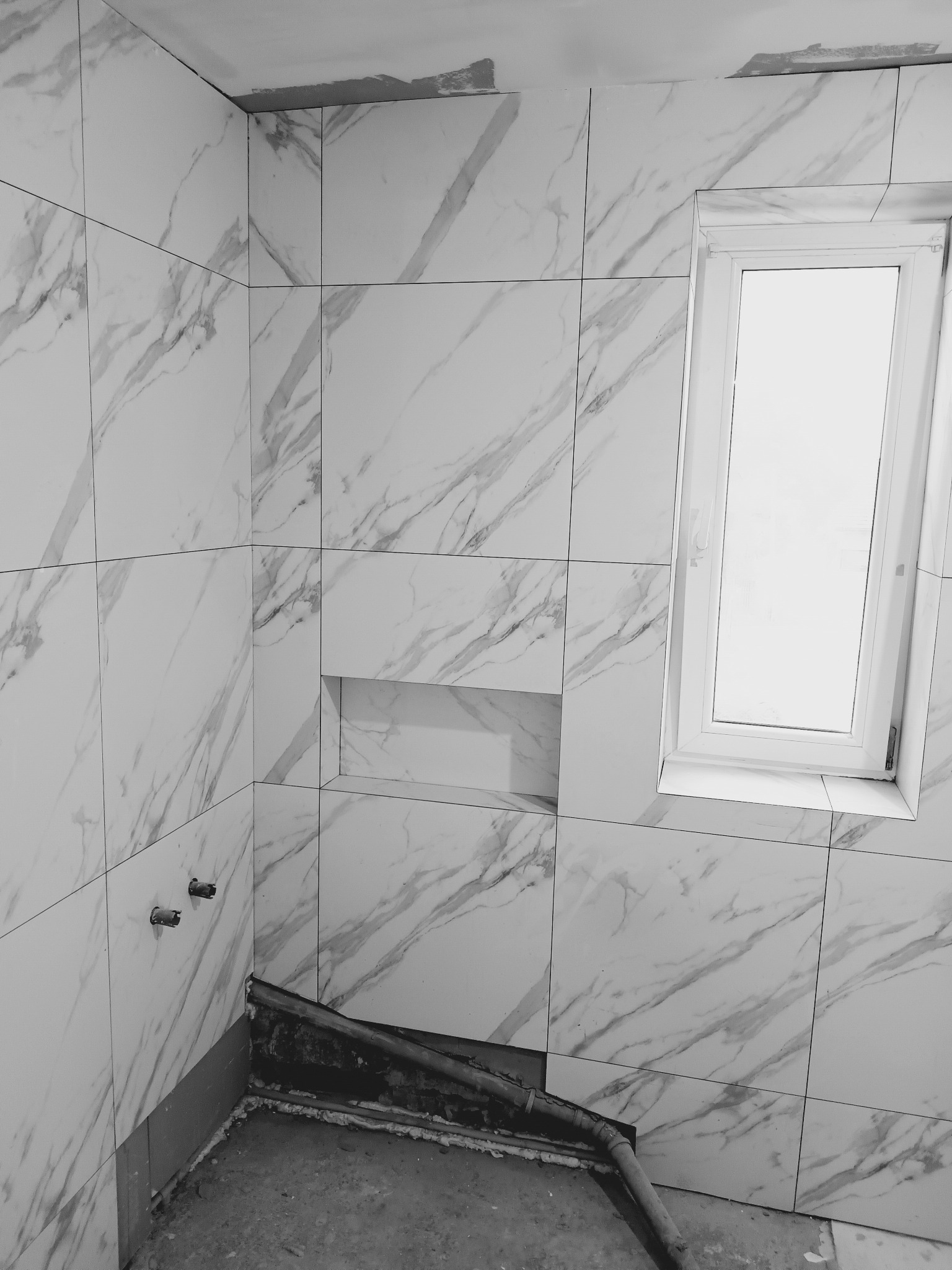 Łazienka w trakcie remontu: białe marmurowe płytki, okno, widoczne rury i przygotowane miejsce na armaturę. Etap prac glazurniczych.