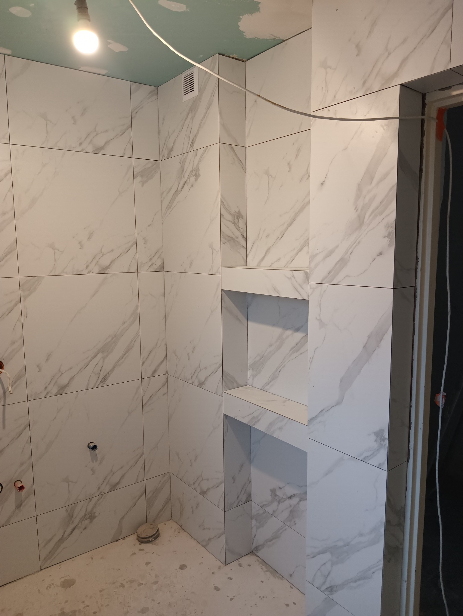 Wykończona łazienka z białymi płytkami imitującymi marmur i wnęką. Widoczny fragment sufitu z zieloną płytą gipsową. Podłoga surowa, przed położeniem posadzki.