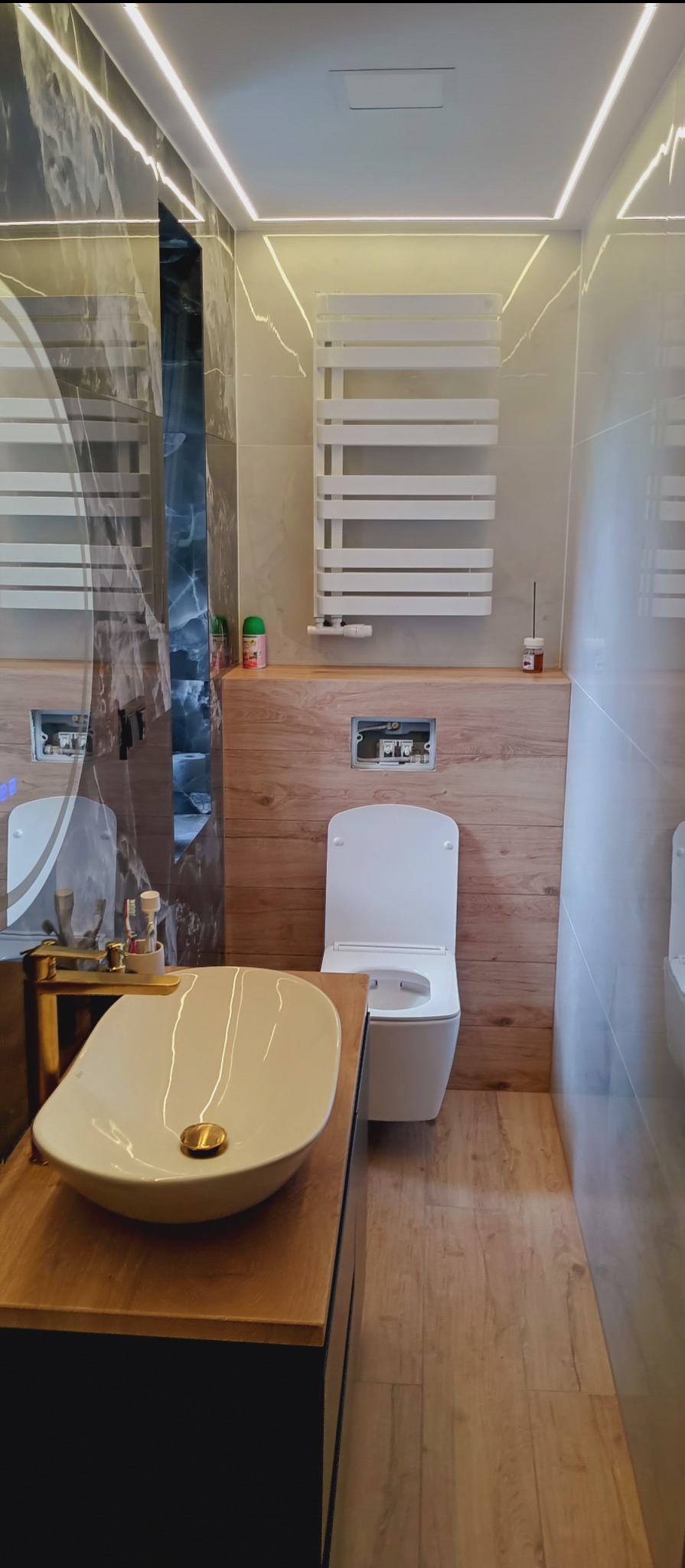 Nowoczesna łazienka z owalną umywalką, złotą armaturą i podświetleniem LED. Na ścianach płytki imitujące marmur i drewno. Grzejnik drabinkowy.