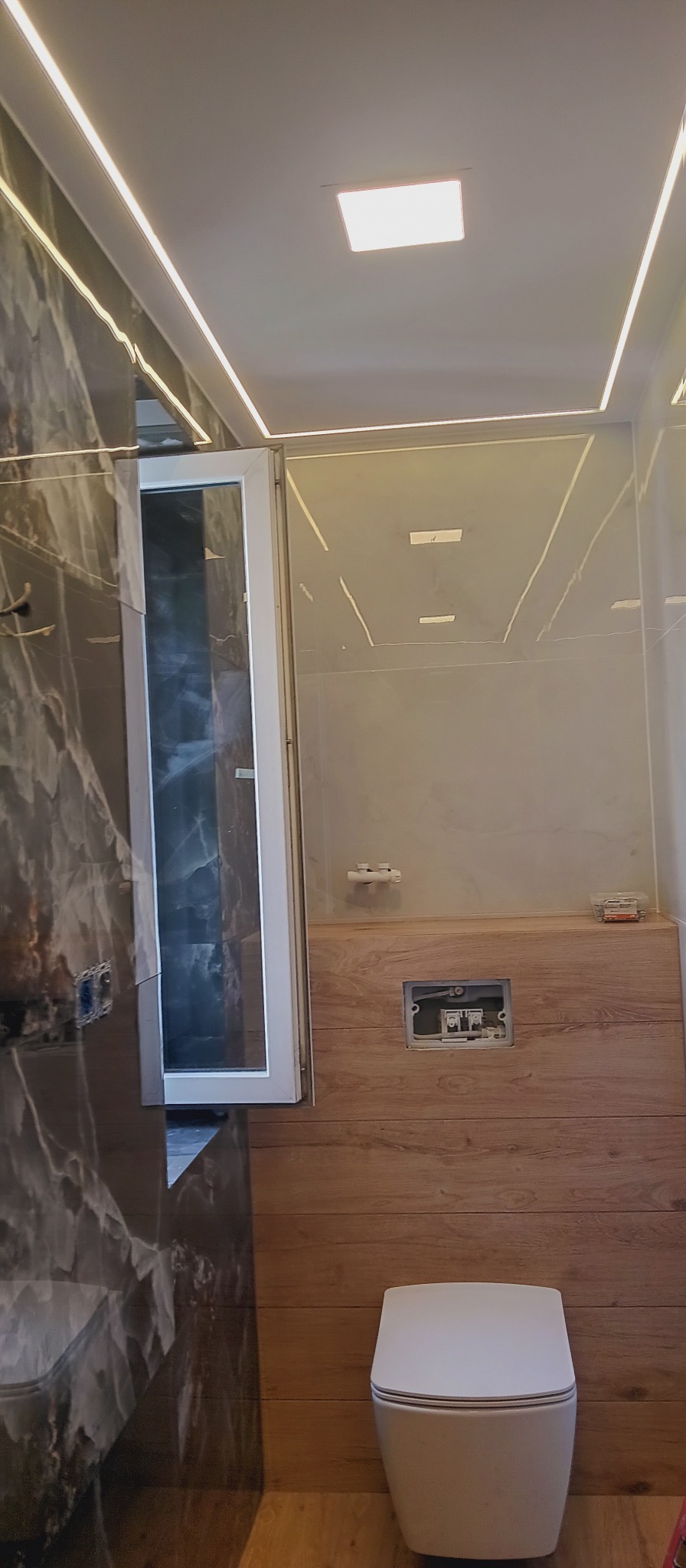 Elegancka łazienka po remoncie: marmurowe i drewnopodobne płytki, podwieszana toaleta, oświetlenie LED. Widoczna otwarta kabina prysznicowa.