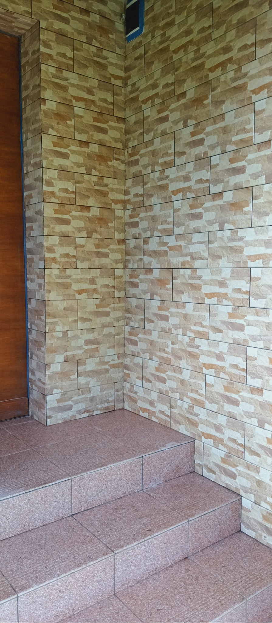 Wykończona ściana zewnętrzna z dekoracyjnej, beżowo-brązowej cegły, z widocznymi schodami wejściowymi wyłożonymi płytkami w podobnej kolorystyce. Narożnik budynku.