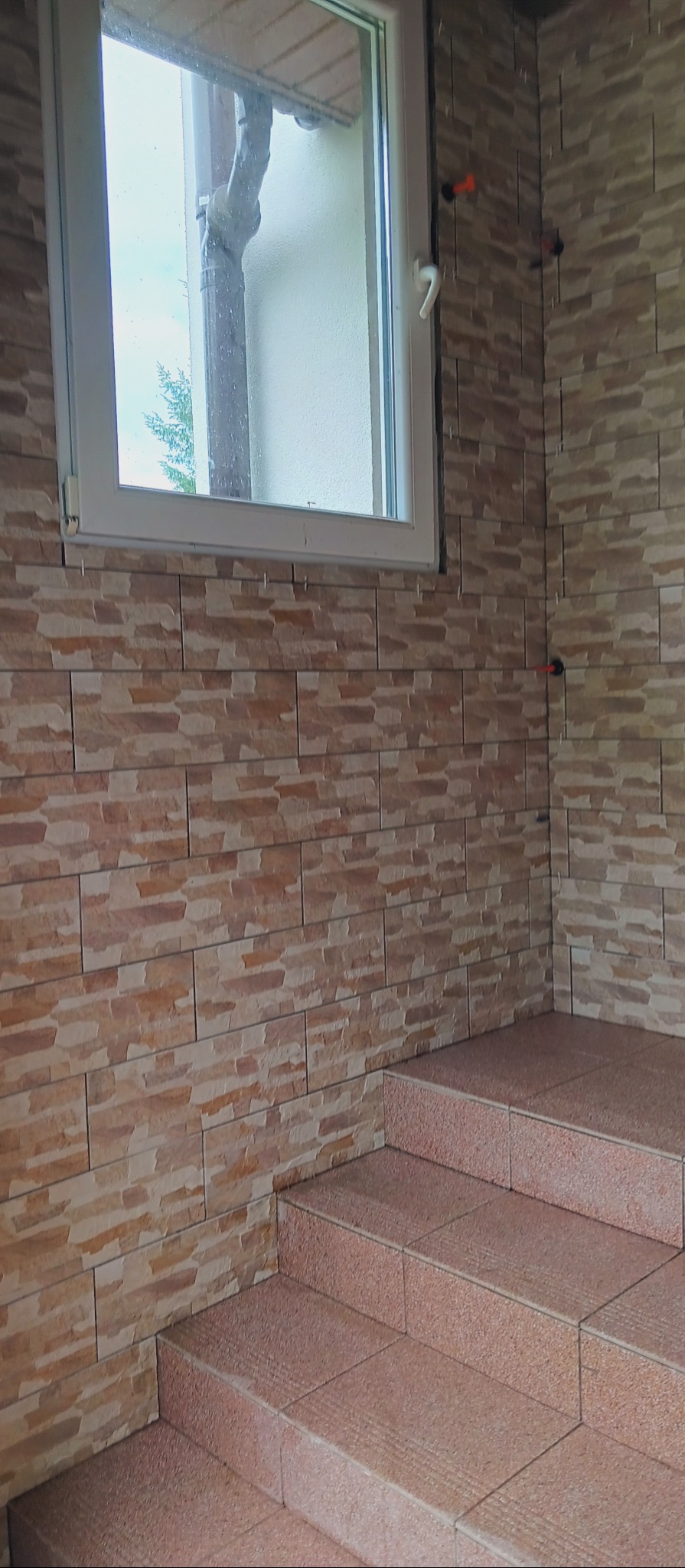 Wykończone schody i ściana z płytek imitujących cegłę, widoczne plastikowe krzyżyki dystansowe. Okno z widokiem na zewnątrz. Wykończenie w odcieniach beżu i brązu.