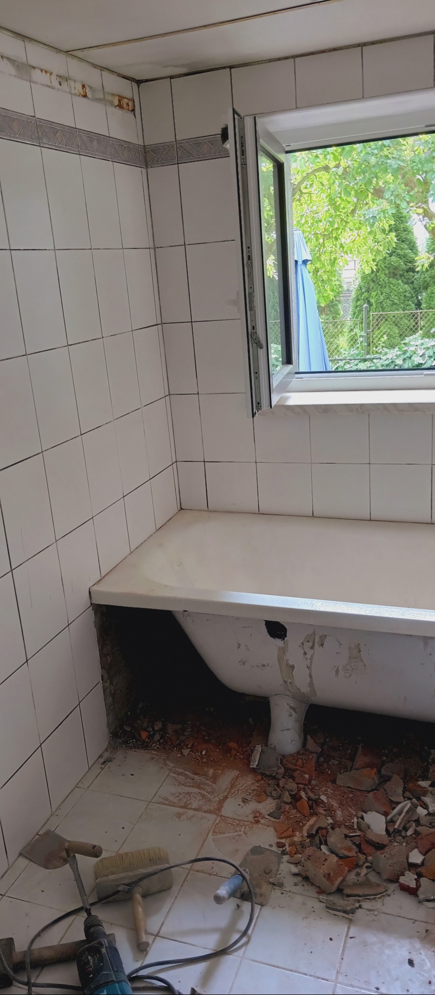 Łazienka w trakcie remontu. Widoczne białe płytki, wanna, okno z widokiem na zieleń, gruz i narzędzia: szpachelki, wiertarka. Częściowo usunięte płytki ścienne.