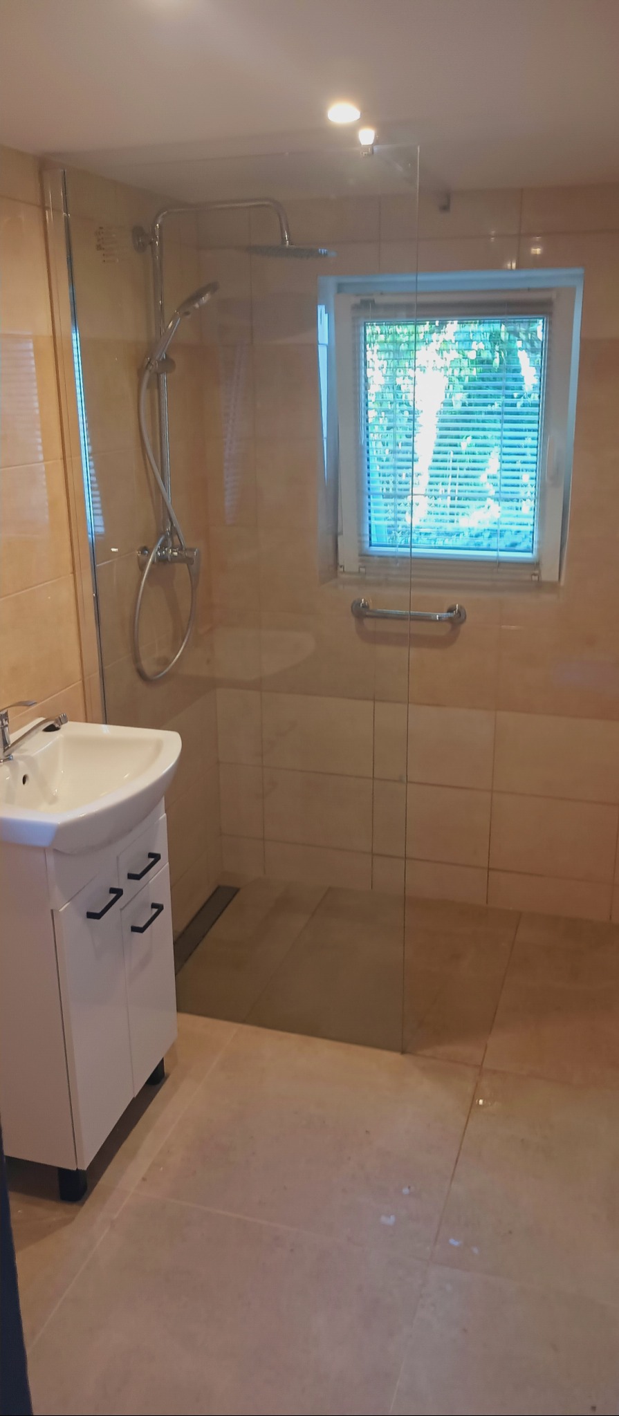 Nowoczesna łazienka po remoncie: kabina prysznicowa walk-in ze szklaną ścianą, biała szafka pod umywalką z czarnymi uchwytami i beżowe płytki na ścianach i podłodze.