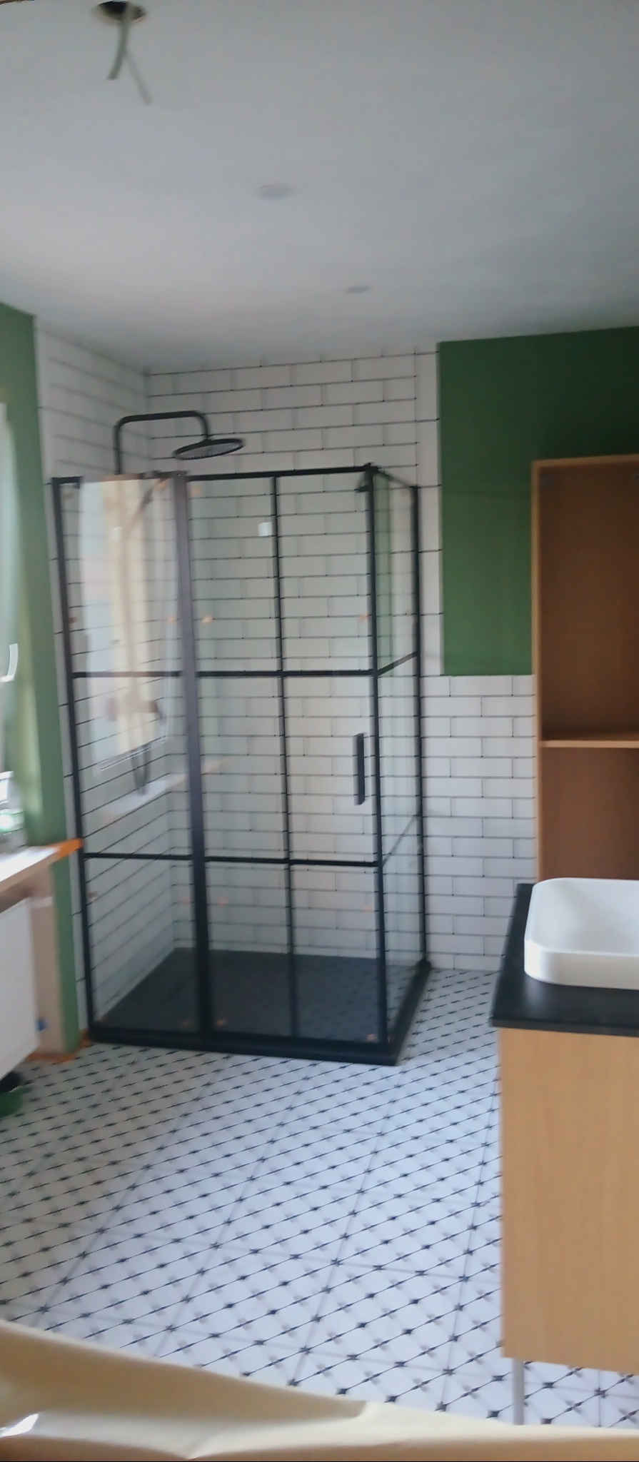 Nowoczesna kabina prysznicowa z czarnymi profilami, białe płytki imitujące cegłę na ścianach, wzorzysta podłoga w wyremontowanej łazience z zielonym akcentem.