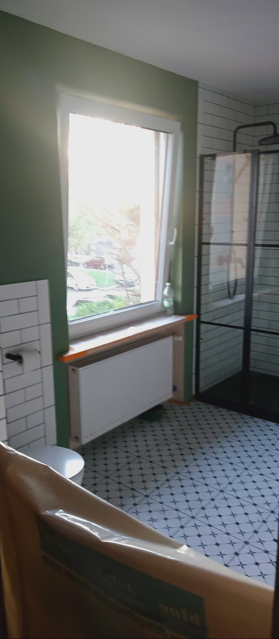 Wykończona łazienka z białą armaturą, grzejnikiem pod oknem i kabiną prysznicową w stylu retro. Płytki podłogowe wzorzyste, ściany w białej cegiełce. Zielona ściana.