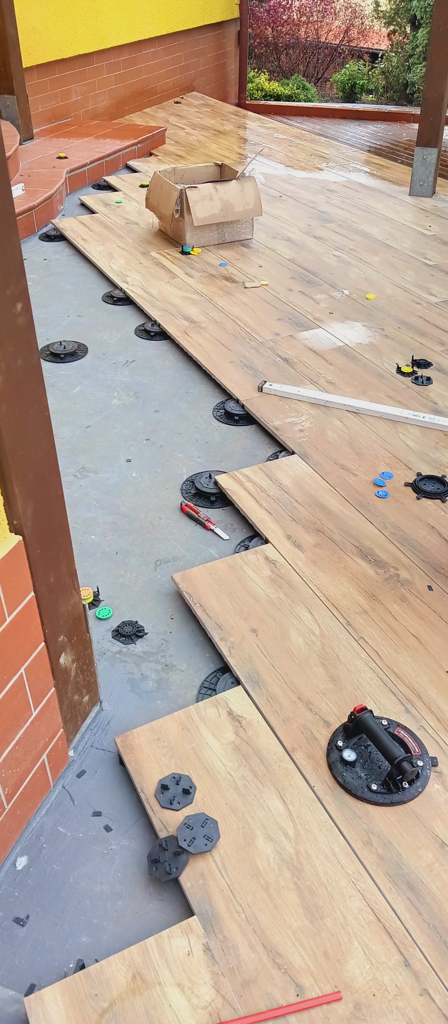 Montaż desek tarasowych na regulowanych wspornikach. Widoczny nóż, przyssawka do przenoszenia płytek i karton. Ujęcie z góry, częściowo ukończony taras.