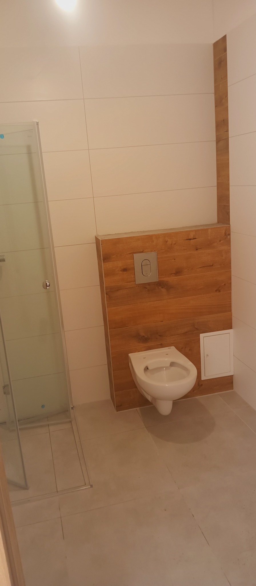 Nowoczesna łazienka z podwieszaną toaletą na drewnianej ściance, białe płytki i szklana kabina prysznicowa. Minimalistyczny design, ciepłe oświetlenie.