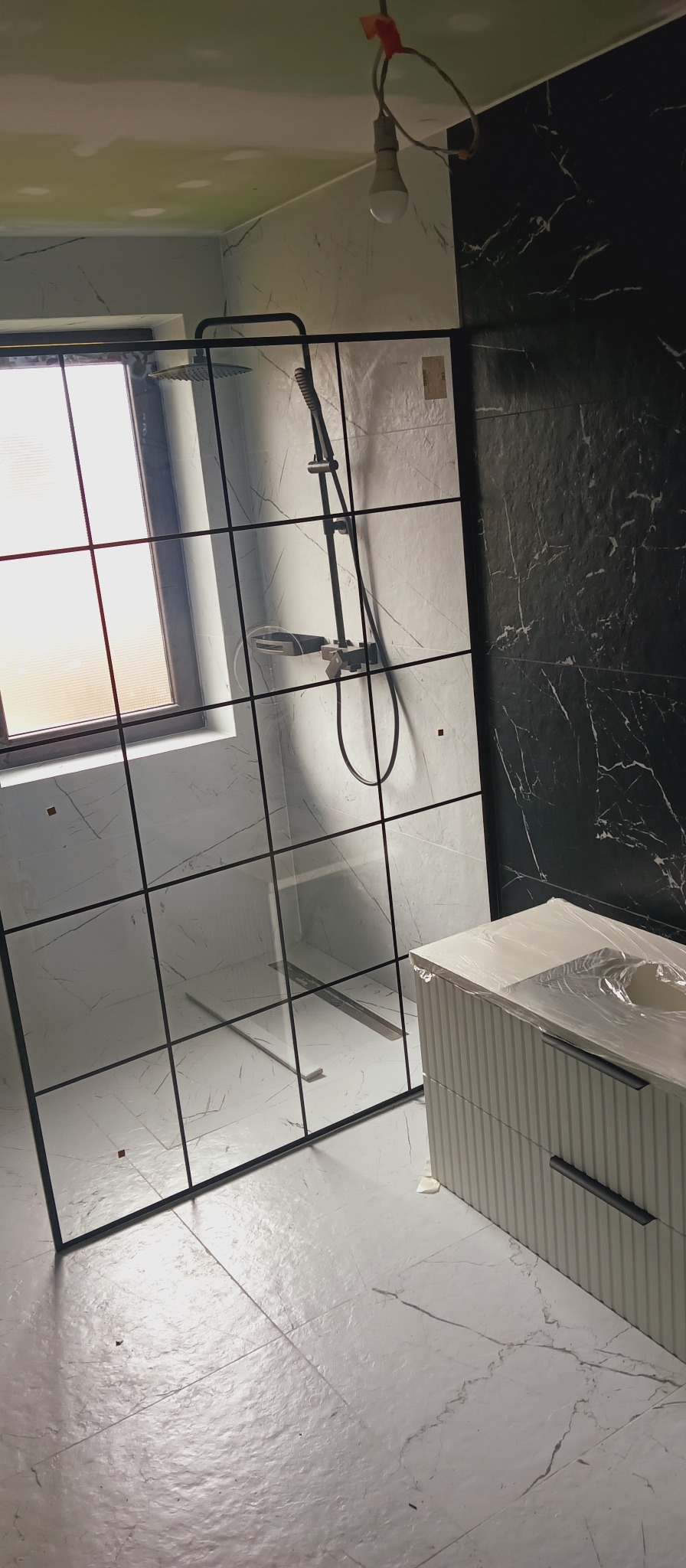 Nowoczesna kabina prysznicowa z czarną ramą na tle marmurowych płytek w łazience. Umywalka z szafką częściowo zakryta folią ochronną. Widok na okno.