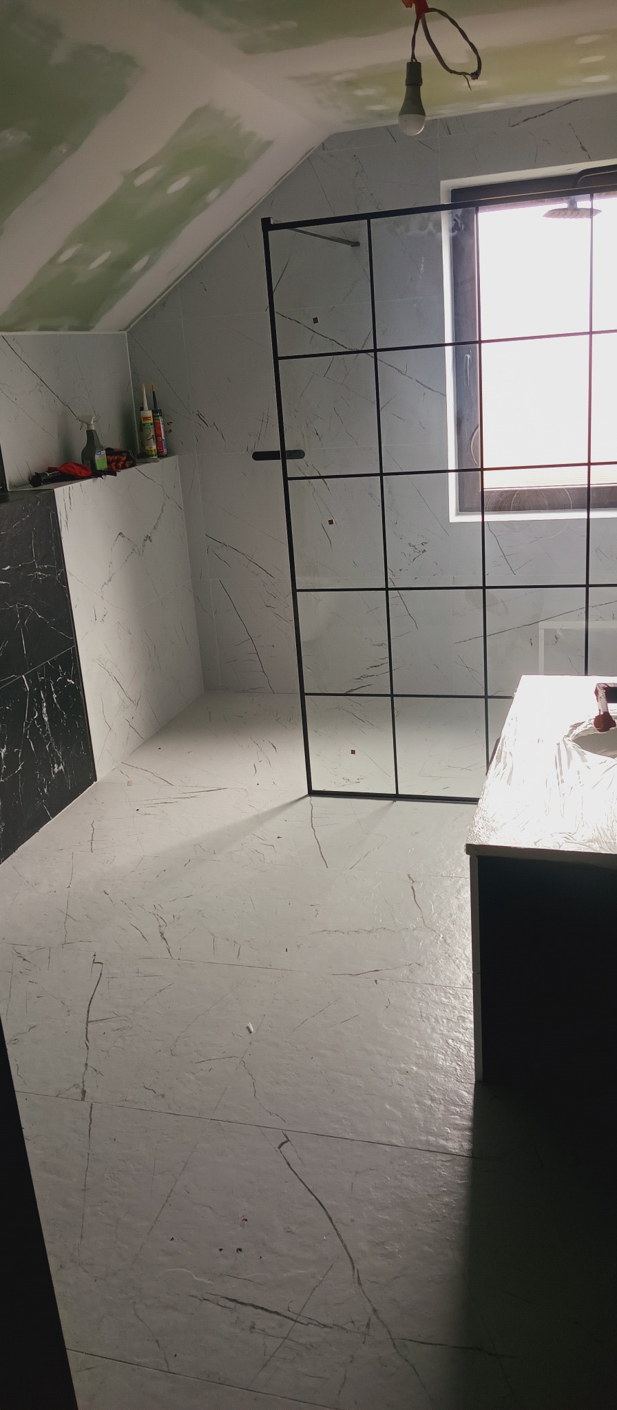 Wykończona łazienka na poddaszu ze ścianami i podłogą w marmurze, kabiną prysznicową z czarnymi profilami i oknem. Widoczne narzędzia i materiały budowlane.