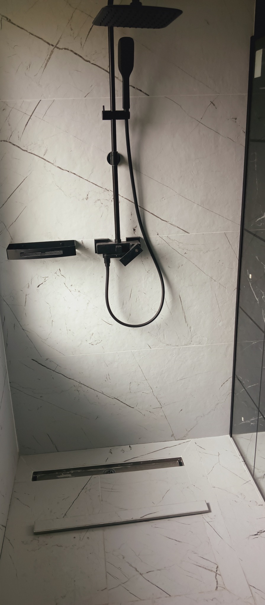 Wykończona łazienka z czarną armaturą: deszczownica, bateria i półka. Białe płytki imitujące marmur z szarymi żyłami. Uwagę przykuwa odpływ liniowy w podłodze.