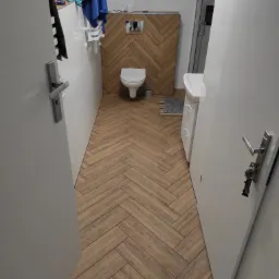 Wnętrze łazienki z podłogą i ścianą za toaletą wyłożoną płytkami imitującymi drewno w jodełkę, biała toaleta i umywalka, widok zza otwartych drzwi.