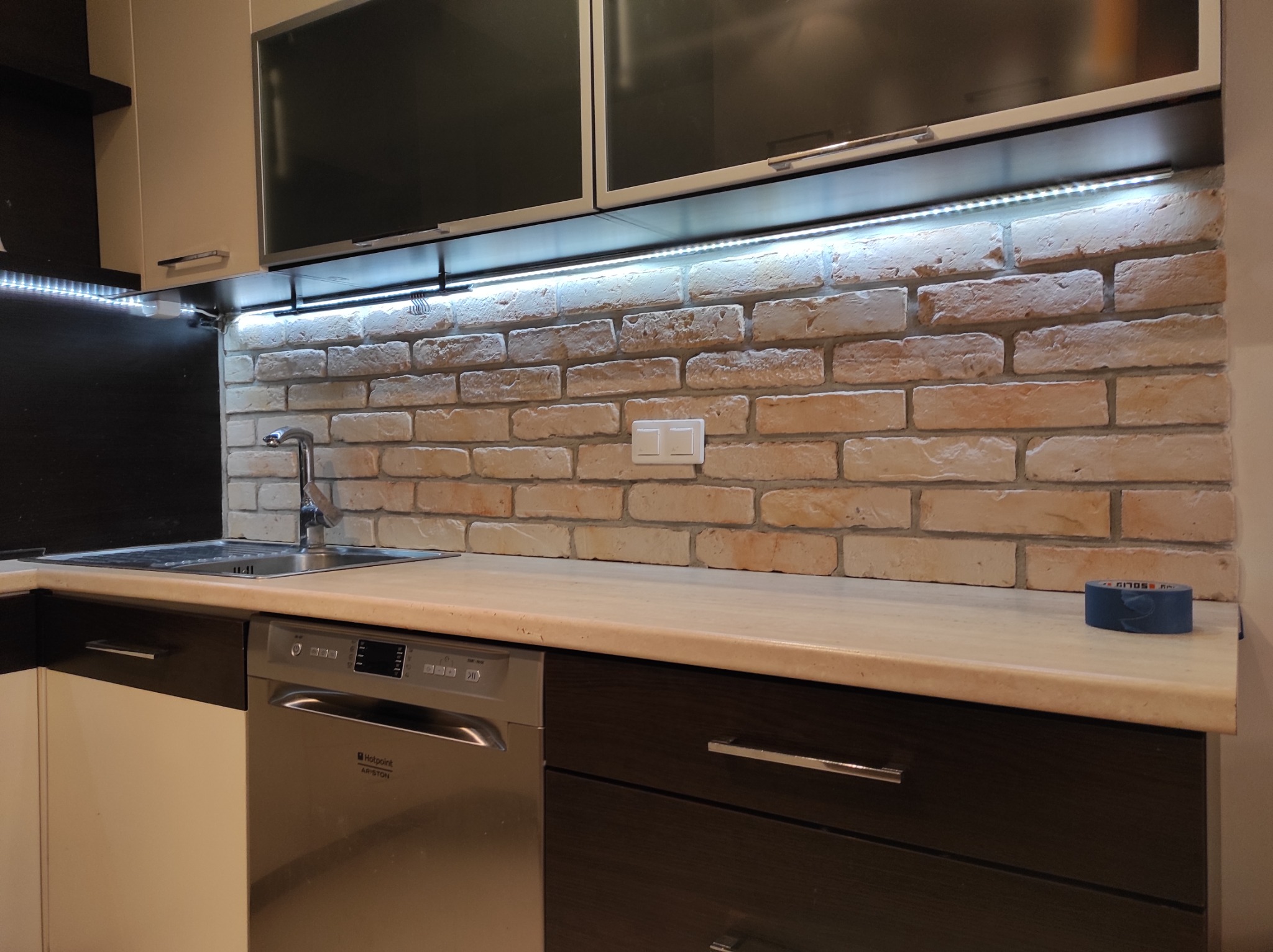 Kuchnia z wykończoną ścianą z cegieł, oświetleniem LED pod szafkami, zlewozmywakiem, baterią i zmywarką Hotpoint Ariston, widoczna taśma malarska na blacie.