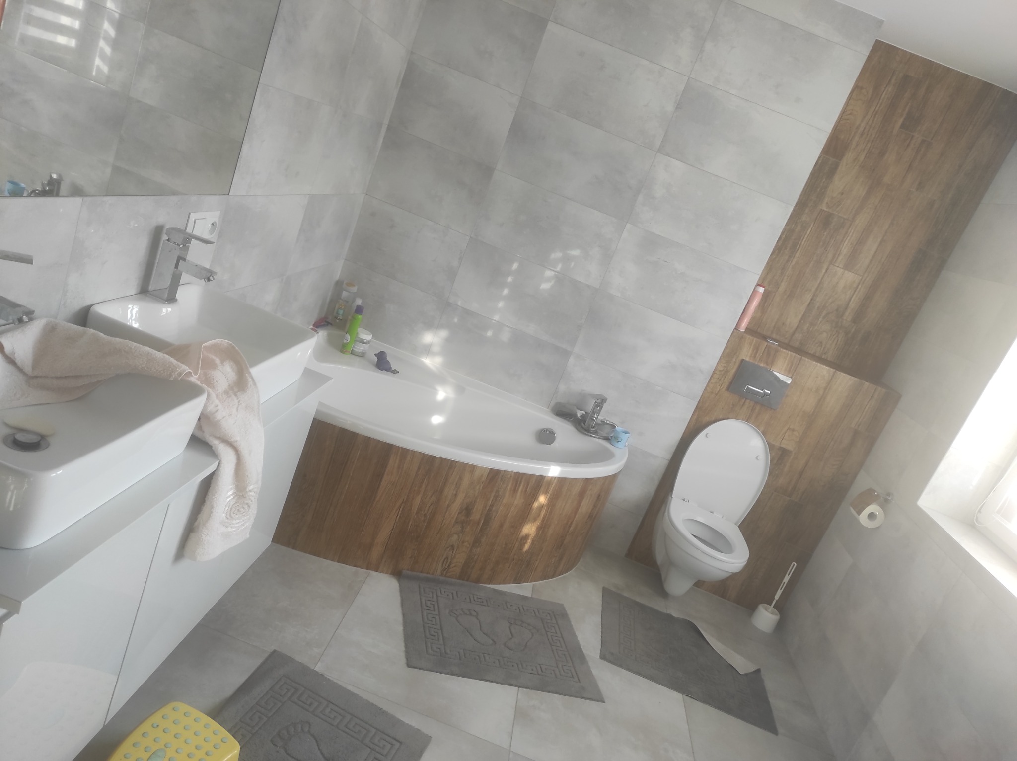 Nowoczesna łazienka z wanną obudowaną drewnem, umywalką nablatową, toaletą i szarymi płytkami imitującymi beton, uzupełniona o dywaniki z motywem stóp.