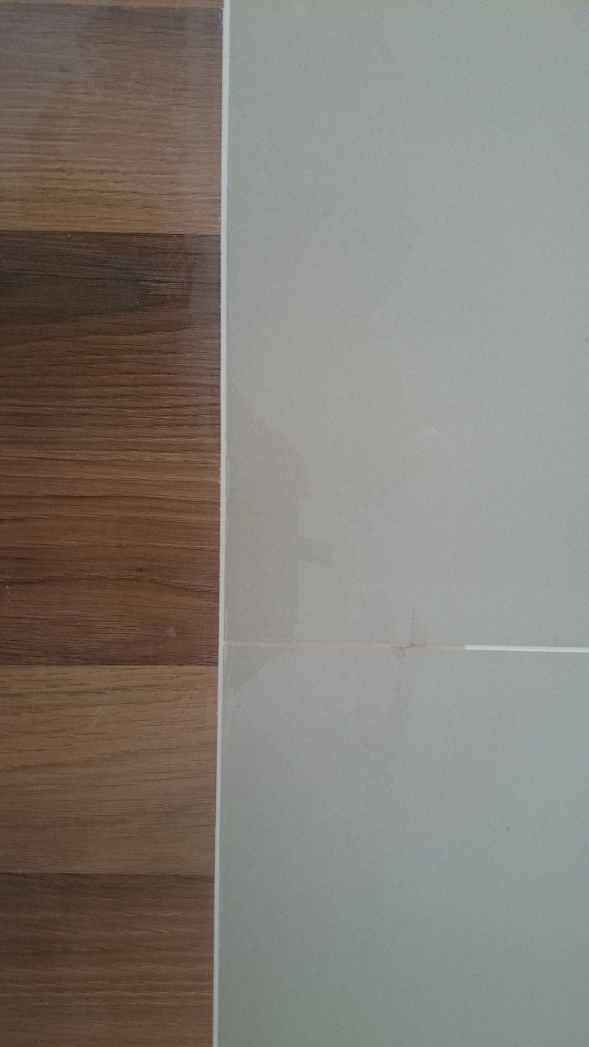 Połączenie brązowych paneli podłogowych z jasnymi, kwadratowymi płytkami ceramicznymi na ścianie, widoczne fugi.