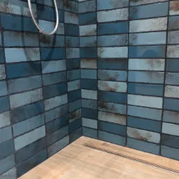 Fragment łazienki z wykończonymi ścianami z niebieskich, cegiełkowych płytek z efektem postarzenia i fragmentem drewnopodobnej podłogi z odpływem liniowym