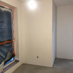 Świeżo pomalowane ściany na biało w pustym pokoju, okno zabezpieczone folią malarską, widoczne gniazdko elektryczne na ścianie.