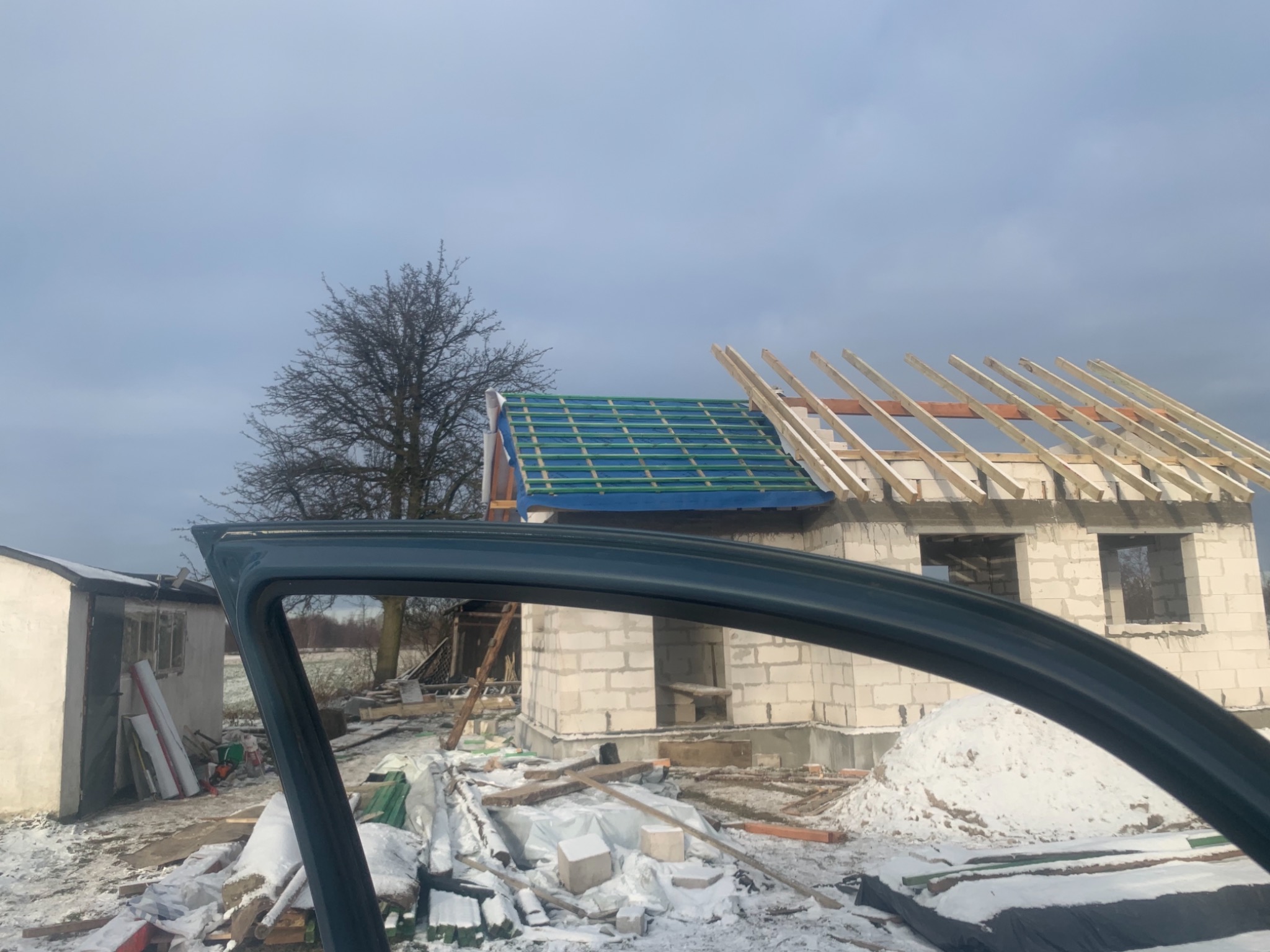 Niedokończony dom z białego bloczka, widoczny przez otwarte okno samochodu, z zielonym zabezpieczeniem na dachu i przygotowaną konstrukcją więźby dachowej, teren wokół ośnieżony.