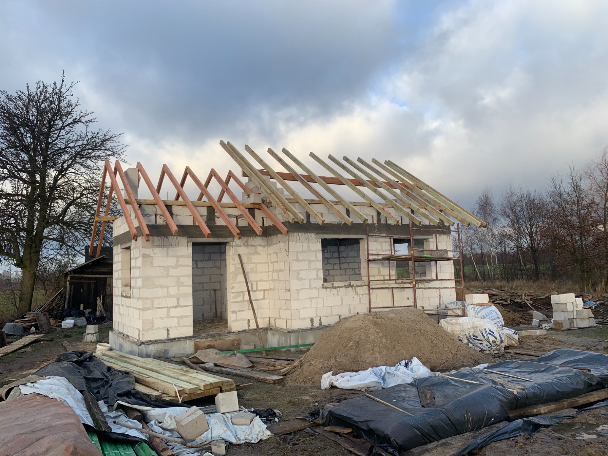 Surowy stan domu z białego bloczka, z częściowo zmontowaną drewnianą konstrukcją dachu, otoczony materiałami budowlanymi i stertą piasku.