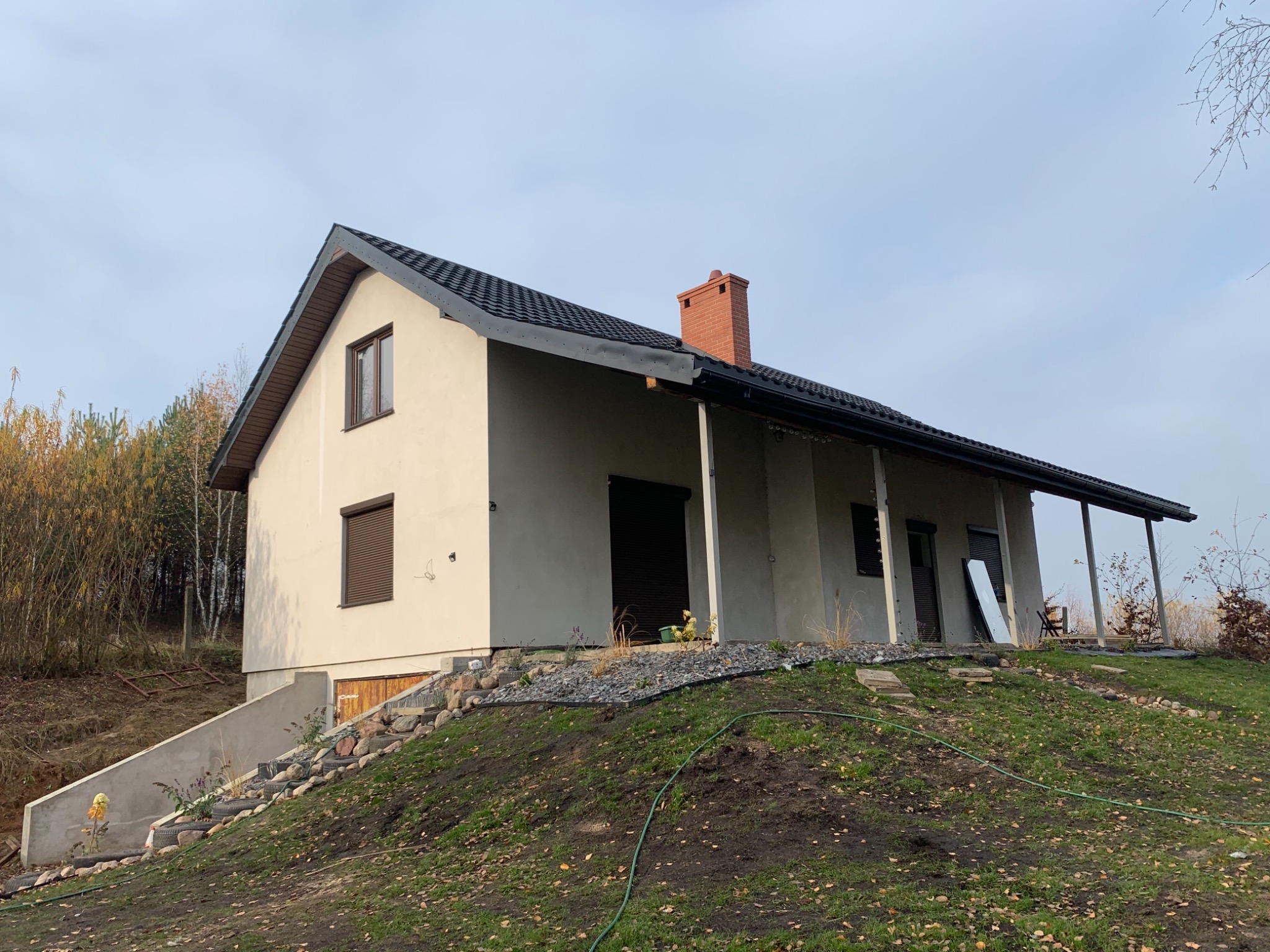 Nowoczesny dom jednorodzinny z szarą elewacją i ciemną dachówką, usytuowany na wzgórzu z kamiennymi schodami i elementami zieleni.