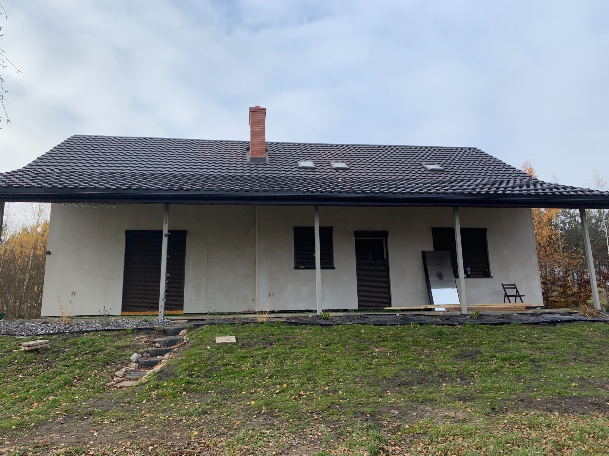 Nowoczesny dom jednorodzinny z ciemną dachówką i ceglanym kominem, elewacja w jasnym odcieniu, zadaszony taras z widocznymi słupami, teren wokół domu w trakcie zagospodarowywania, widoczne schody...