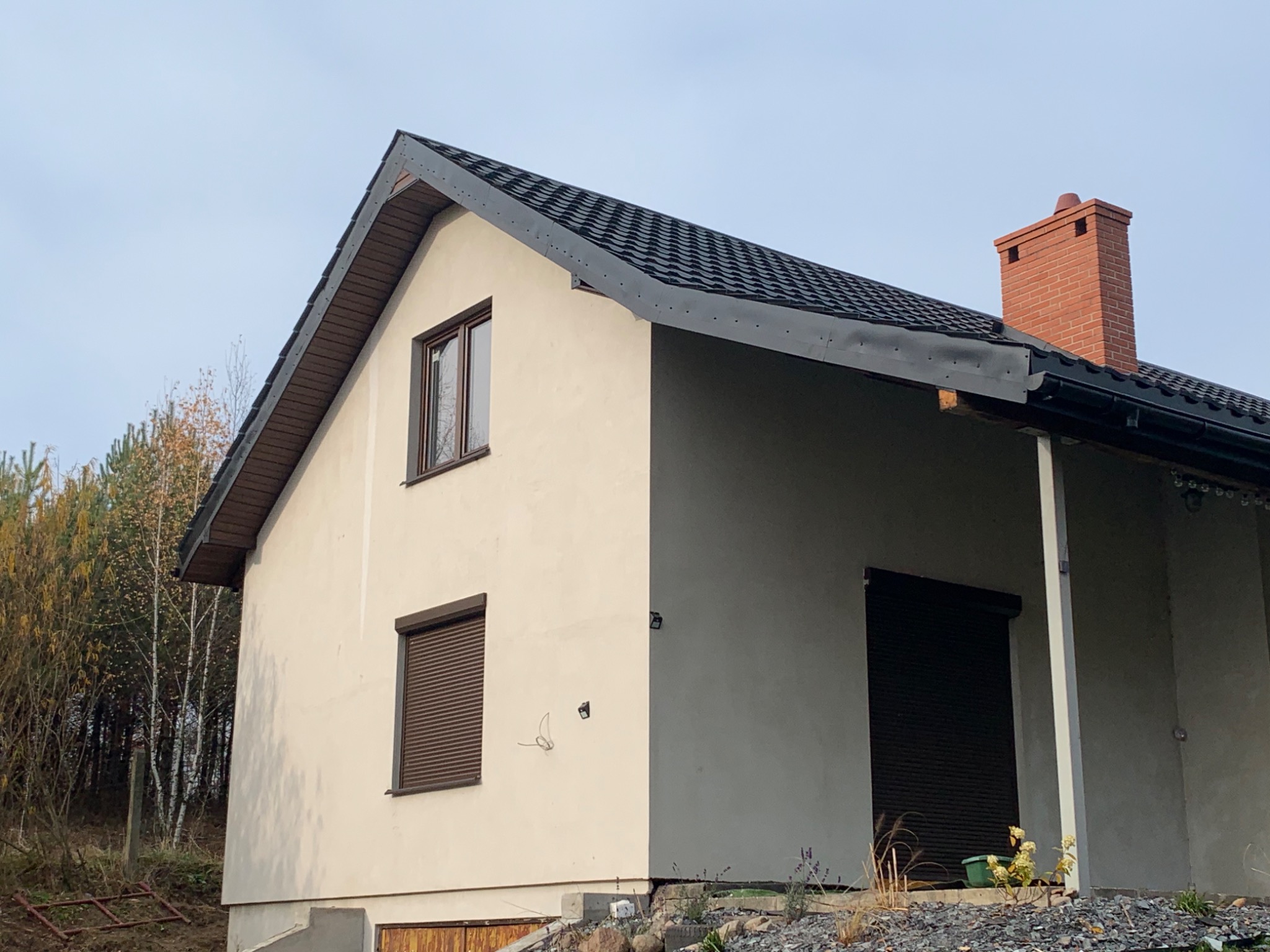 Nowoczesny dom jednorodzinny z beżową elewacją, ciemną dachówką, brązowymi oknami z roletami zewnętrznymi i ceglanym kominem.