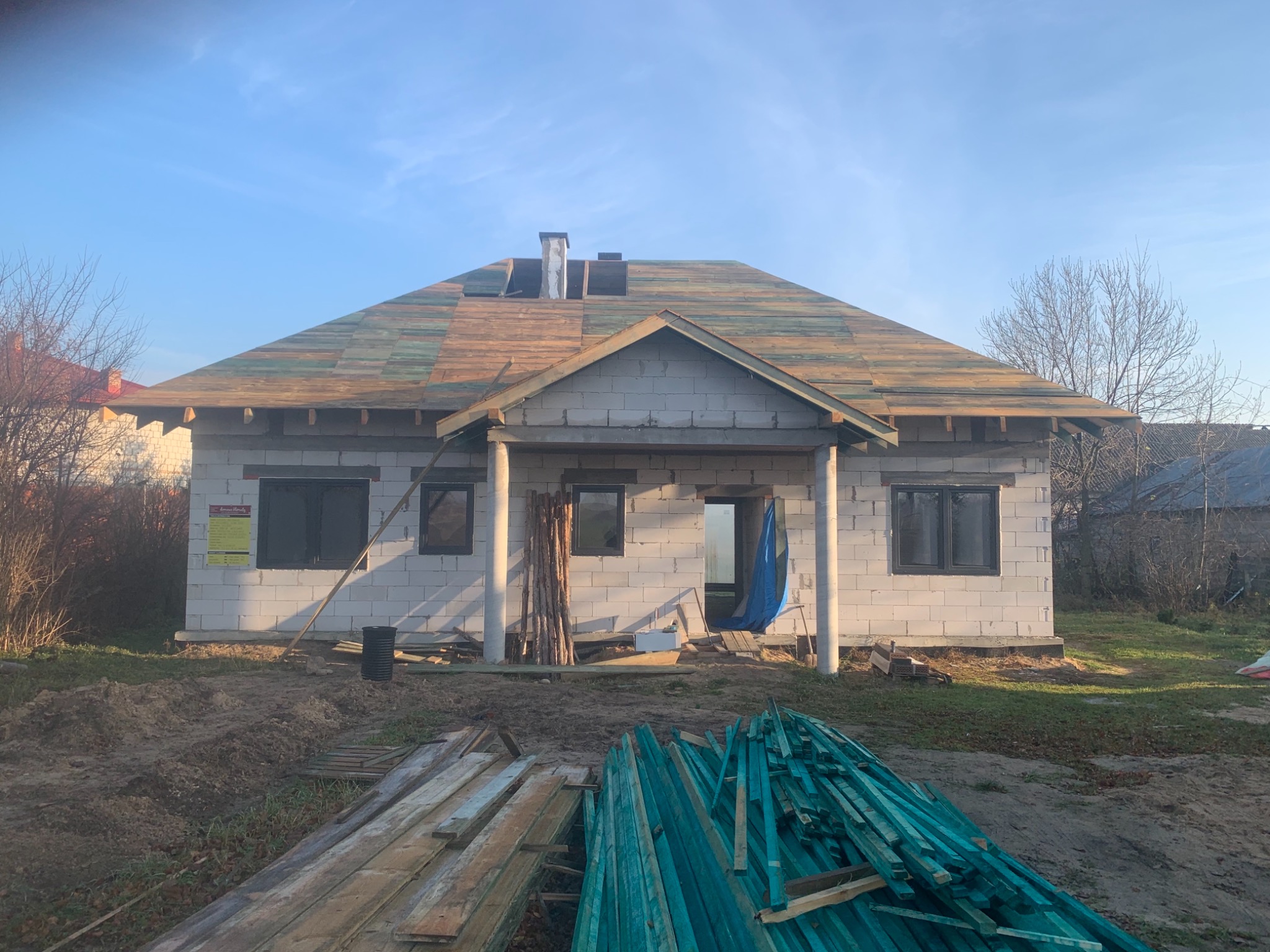 Surowy dom z białego pustaka w trakcie budowy, z częściowo położonym drewnianym dachem, widoczne stosy drewnianych łat na pierwszym planie, jesienne otoczenie.