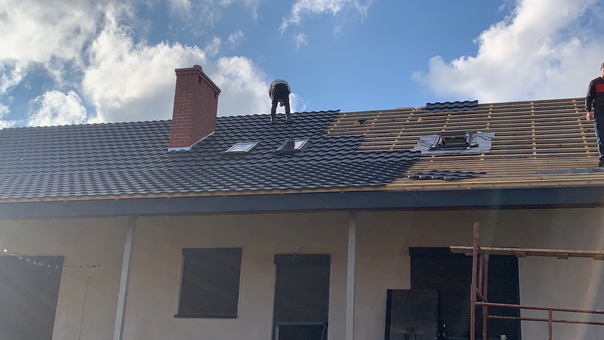Montaż czarnej blachodachówki na dachu domu jednorodzinnego z ceglanym kominem i oknami dachowymi; widoczne elementy konstrukcji dachu oraz pracownicy.
