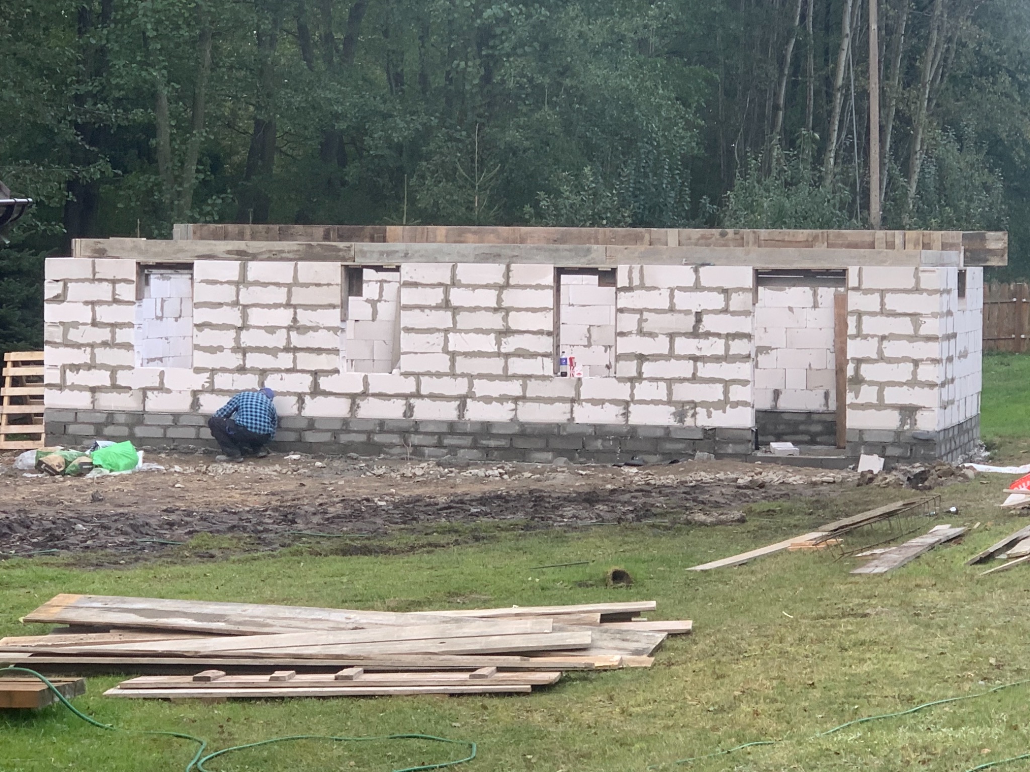 Wznoszenie ścian parterowego budynku z białych bloczków, widoczny murarz w niebieskiej koszuli podczas pracy, w tle zalesiony teren.