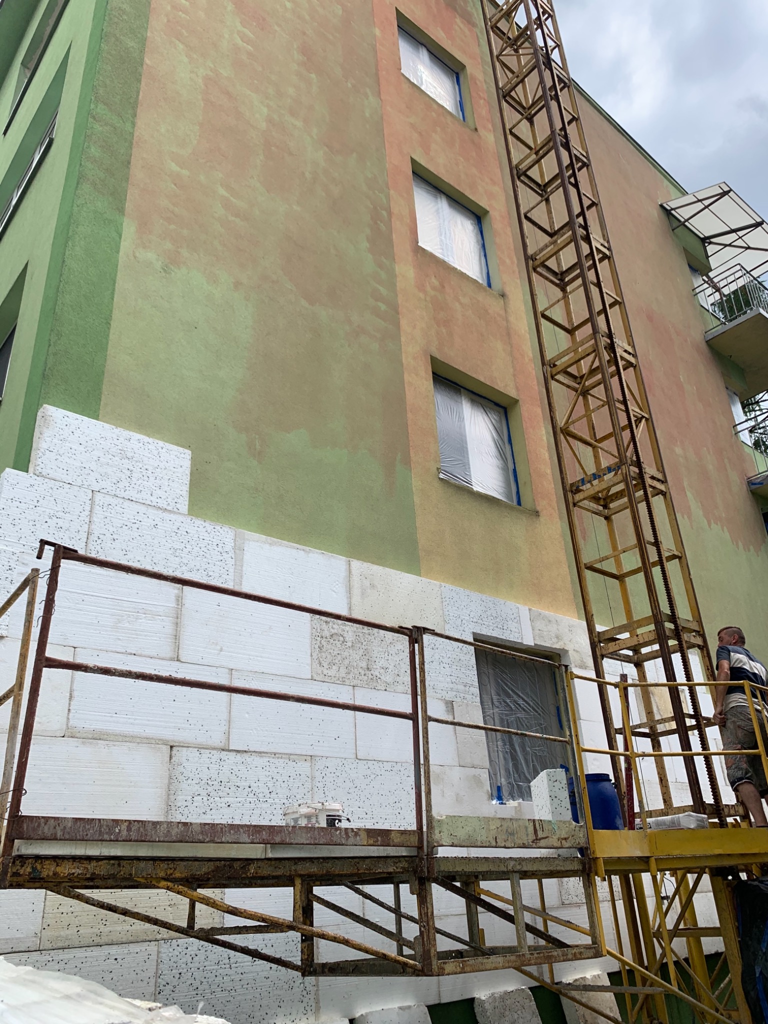 Budynek wielorodzinny w trakcie ocieplania; widoczne białe bloki styropianowe, metalowe rusztowanie i drabina na tle elewacji w odcieniach zieleni i beżu, okna zabezpieczone folią