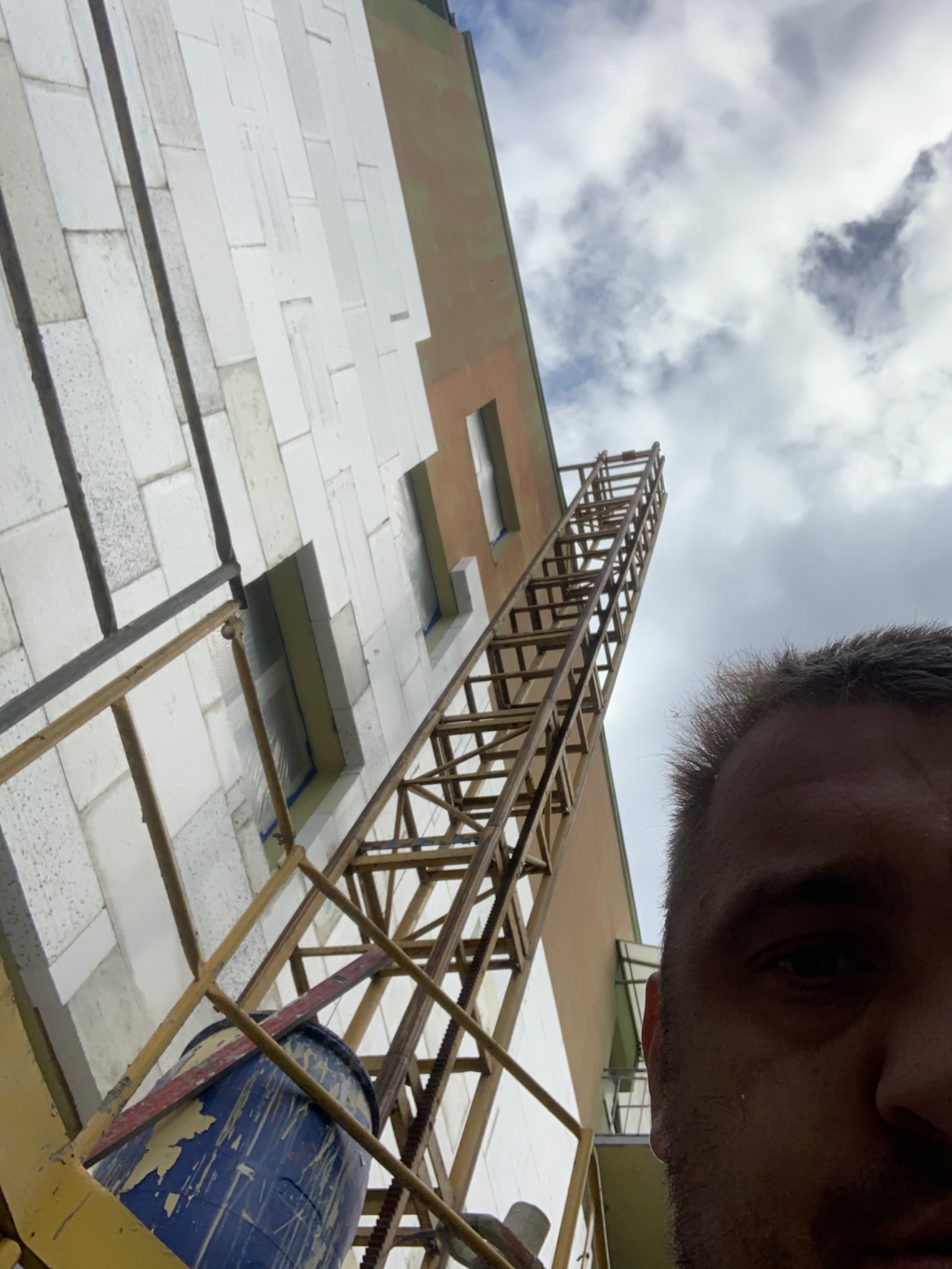 Prace na wysokościach: rusztowanie przy ścianie budynku z ociepleniem z białych płyt styropianowych, widoczny niebieski pojemnik i fragment twarzy osoby fotografującej.