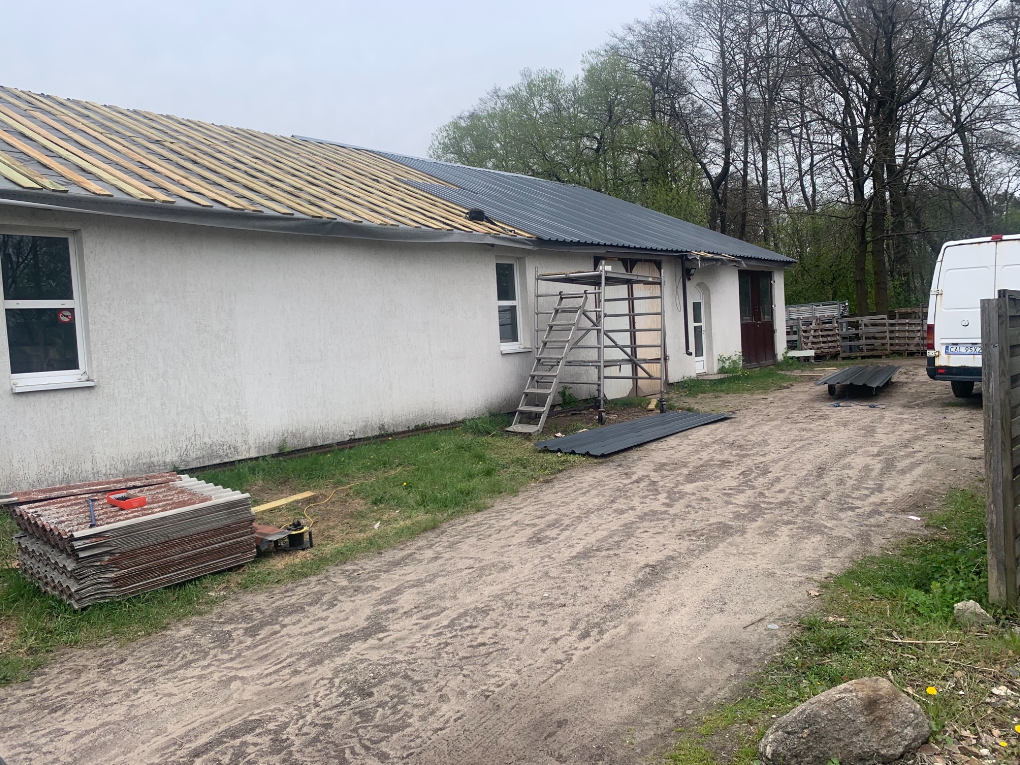 Połowa dachu budynku pokryta szarą blachą trapezową, druga połowa w trakcie montażu łat drewnianych, rusztowanie przy ścianie, sterta blachy falistej na ziemi.