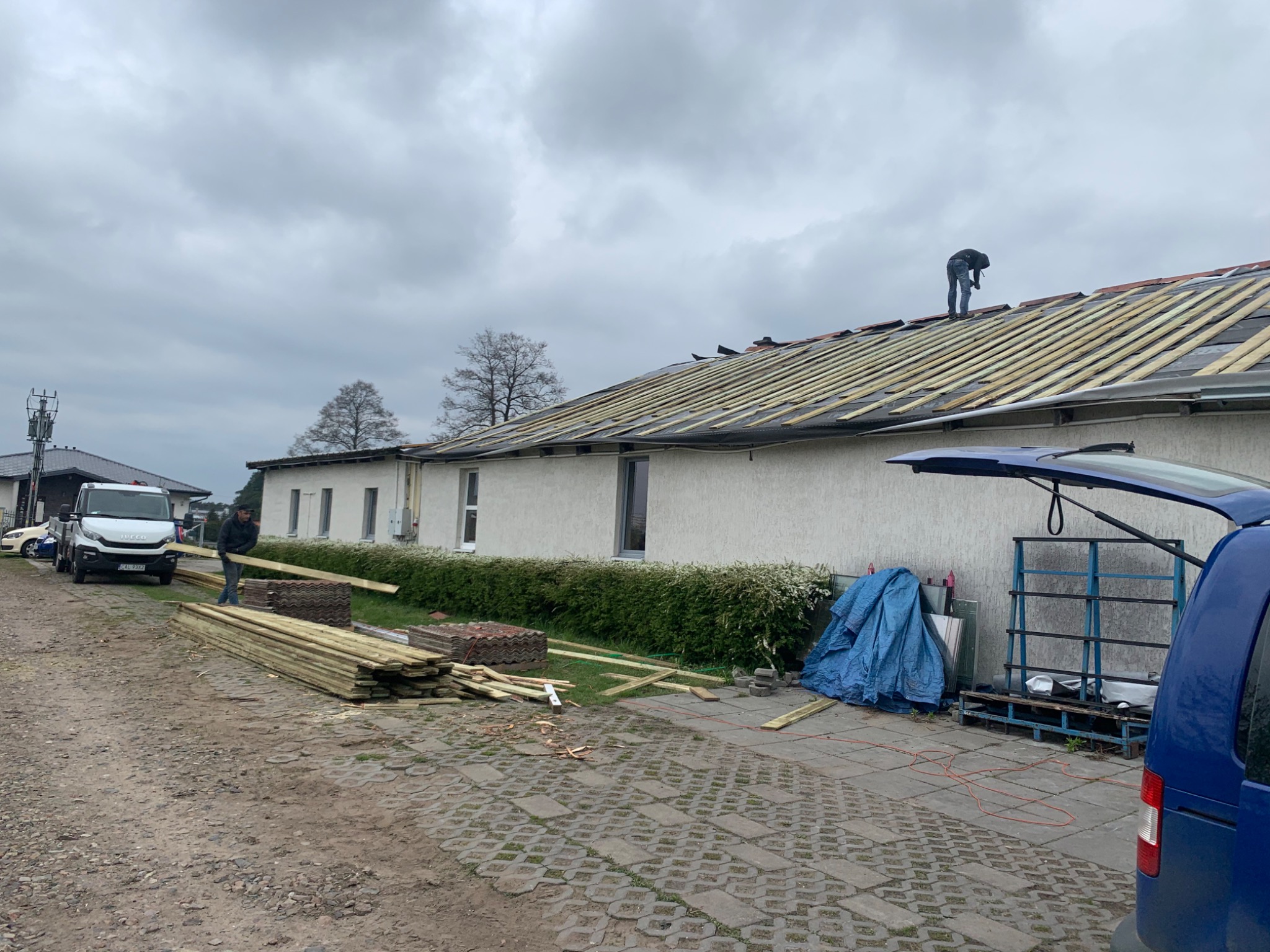 Prace dekarskie: montaż łat na dachu budynku jednokondygnacyjnego, widoczne drewniane elementy, pracownik na dachu, materiały budowlane i samochody dostawcze w tle, pochmurne niebo.