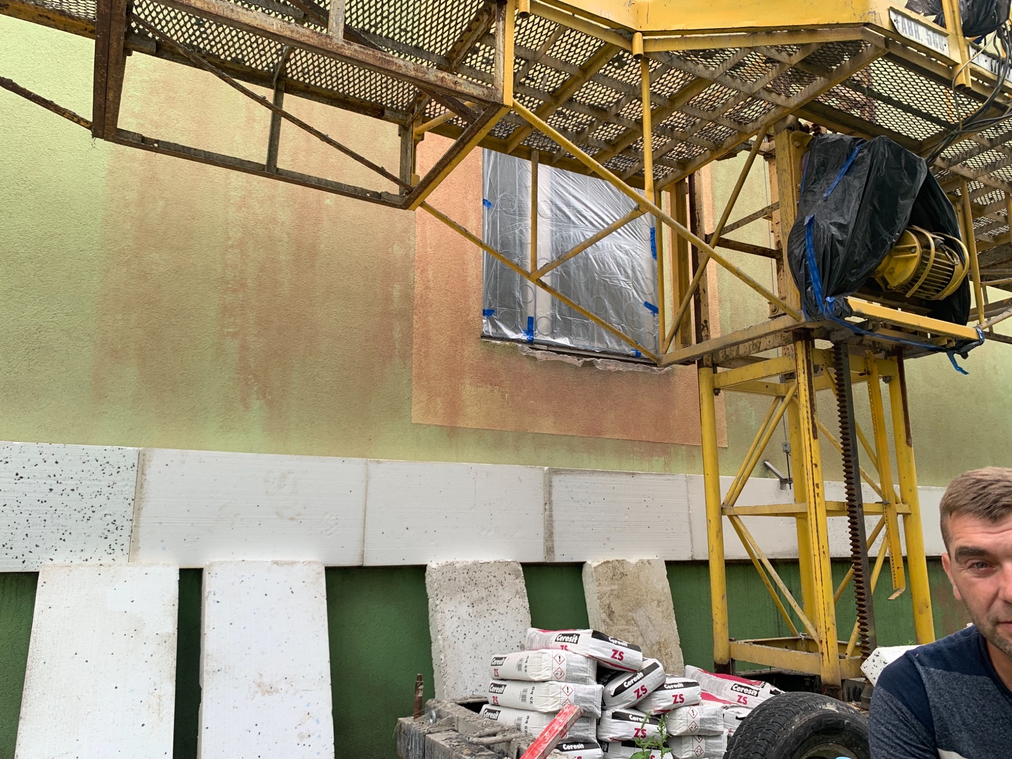 Elewacja budynku w trakcie prac: styropianowe płyty częściowo przyklejone, okno zabezpieczone folią, widoczny dźwig budowlany i worki z zaprawą klejową Ceresit ZS.