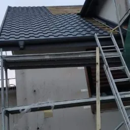 Fragment dachu z nową, ciemną dachówką, widoczne rusztowanie i drabina, część starego poszycia jeszcze nie wymieniona.