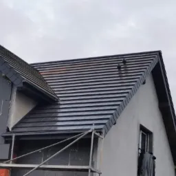 Fragment domu w trakcie prac budowlanych: nowy, ciemny dach z dachówki ceramicznej, widoczne rusztowanie i ocieplenie ścian zewnętrznych styropianem.