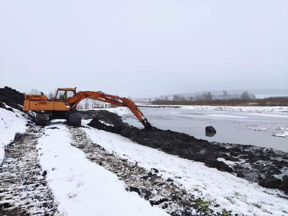 Pomarańczowa koparka Atlas na gąsienicach pogłębia brzeg zbiornika wodnego w zimowej scenerii, śnieg leży na ziemi i częściowo na tafli wody, w tle widoczne drzewa.