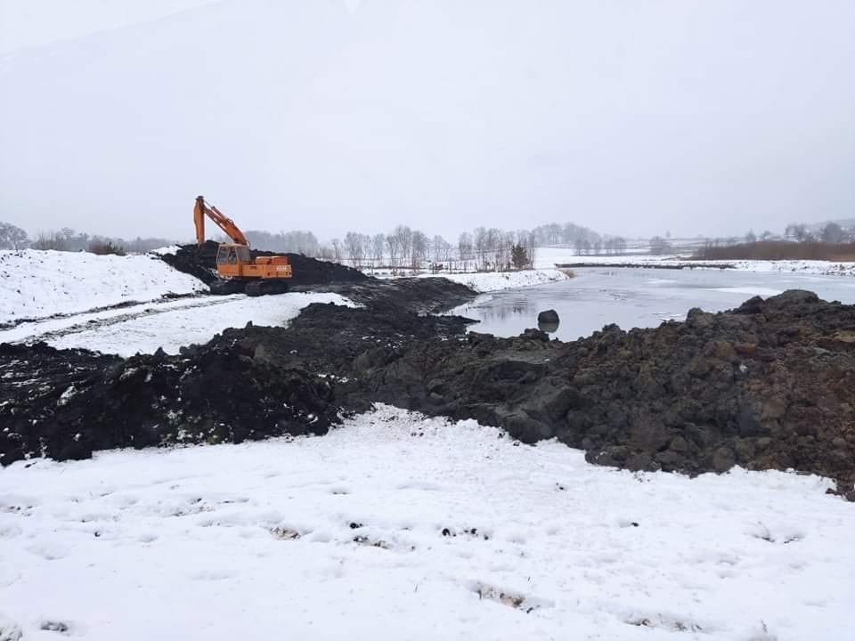 Pomarańczowa koparka pracująca w zimowym krajobrazie przy zbiorniku wodnym, z hałdami czarnej ziemi i śniegiem na pierwszym planie.