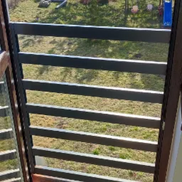 Widok z wnętrza przez otwarte okno z zewnętrznymi żaluzjami fasadowymi, na zewnątrz trawnik z huśtawką i drzewami, na podłodze narzędzia i karton zabezpieczający.