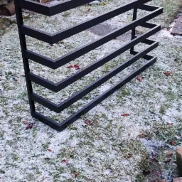 Czarna, metalowa konstrukcja spawana o geometrycznym kształcie, przypominająca schody lub drabinę, ustawiona na trawie pokrytej szronem w ogrodzie.