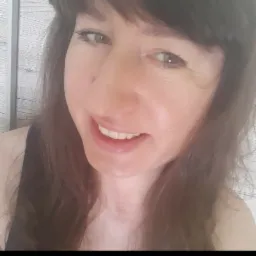 Portret uśmiechniętej kobiety z brązowymi włosami i zielonymi oczami, na pierwszym planie widoczne białe zęby, tło stanowią pionowe panele ścienne o subtelnej fakturze.