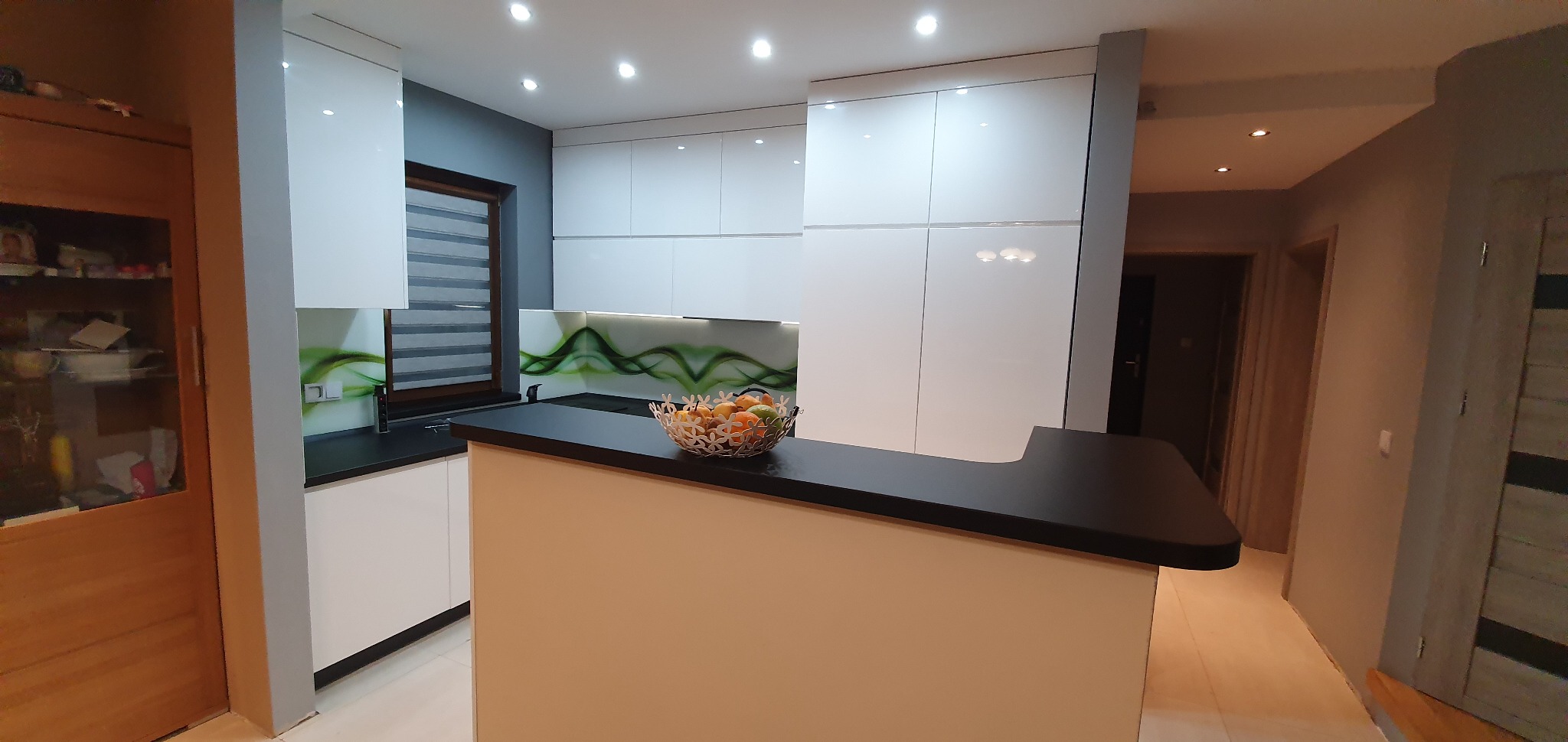 Nowoczesna, biała kuchnia z czarnym blatem w Lubinie, z dekoracyjnym panelem szklanym w zielone fale i owocami w ozdobnej misie na półwyspie.