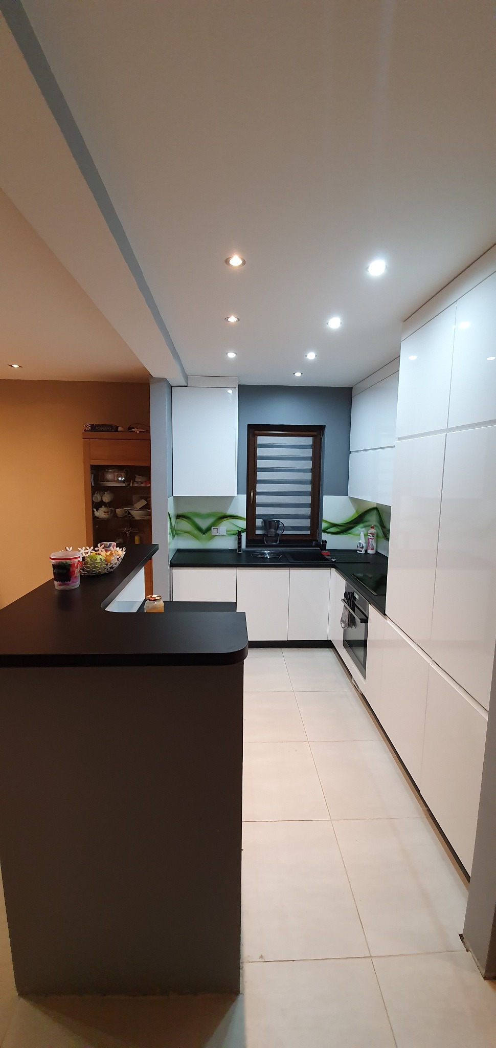 Nowoczesna kuchnia z białymi frontami szafek, czarnym blatem i barkiem, wzorzystą zieloną ścianą nad blatem oraz oknem z żaluzjami poziomymi.