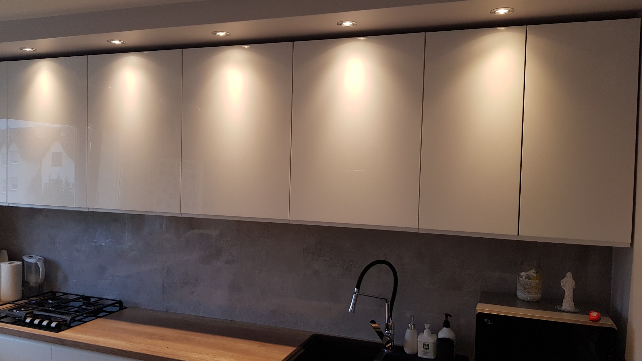 Białe, lakierowane szafki kuchenne podświetlone punktowym oświetleniem sufitowym, z blatem z drewnopodobnego materiału i szarą ścianą nad blatem.
