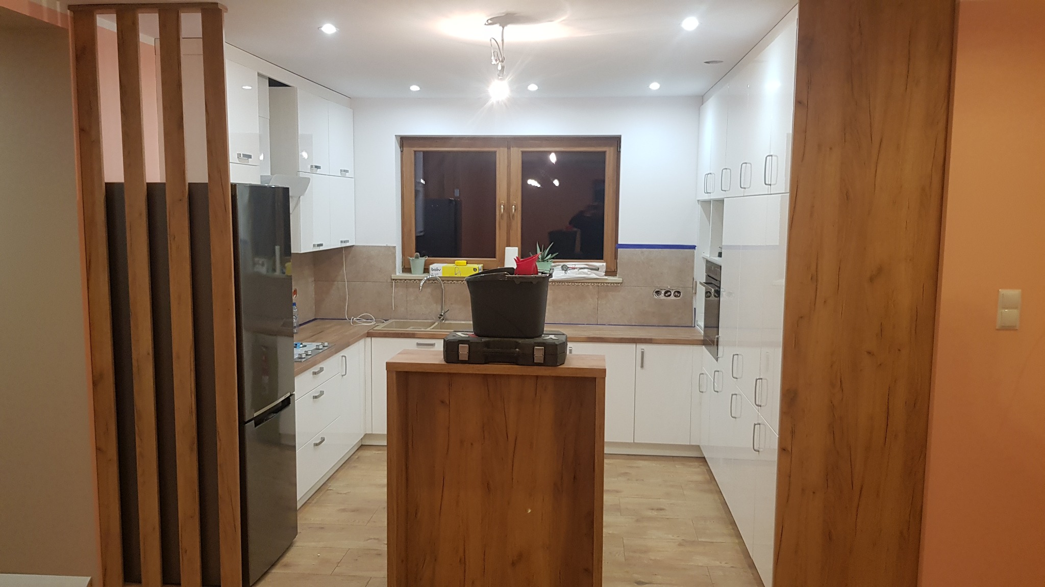 Wnętrze kuchni w trakcie remontu: białe szafki kuchenne, drewniany panel dekoracyjny, okno z widokiem na zewnątrz, na wyspie kuchennej stoi wiadro i skrzynka narzędziowa, zabezpieczone ściany taśmą...