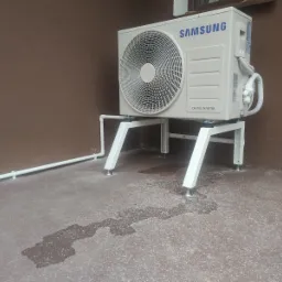 Biała jednostka zewnętrzna klimatyzatora marki Samsung, zamontowana na podwyższonym, metalowym stelażu, z widocznymi śladami wilgoci na podłożu i rurą odprowadzającą skropliny biegnącą wzdłuż...