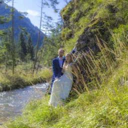 Para młoda w plenerze: on w granatowym garniturze, ona w zwiewnej sukni, pozują na tle górskiego potoku i skał porośniętych trawą, w słoneczny dzień.