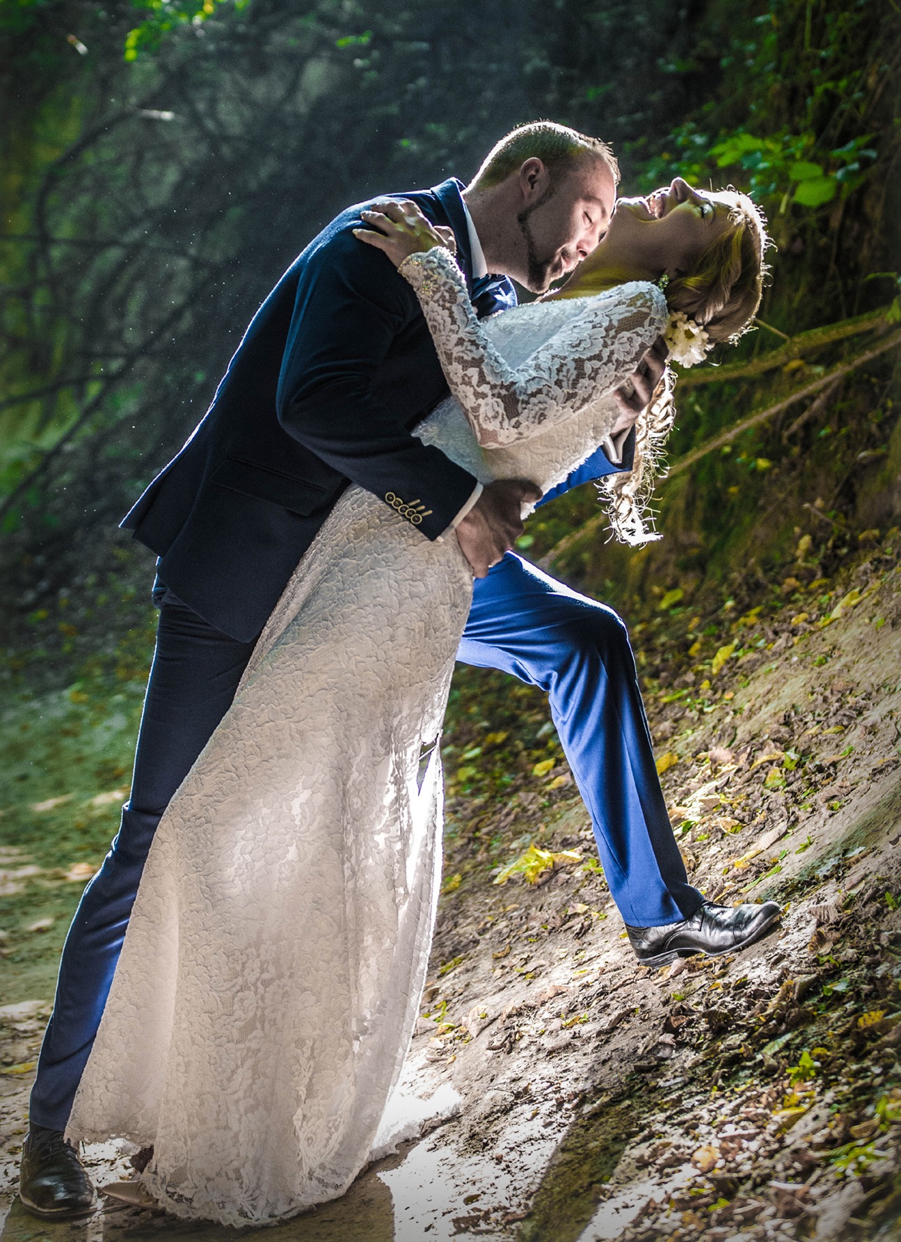 Para młoda w tańcu na leśnej ścieżce. Panna młoda w koronkowej sukni, pan młody w granatowym garniturze. Romantyczna sceneria, miękkie światło.
