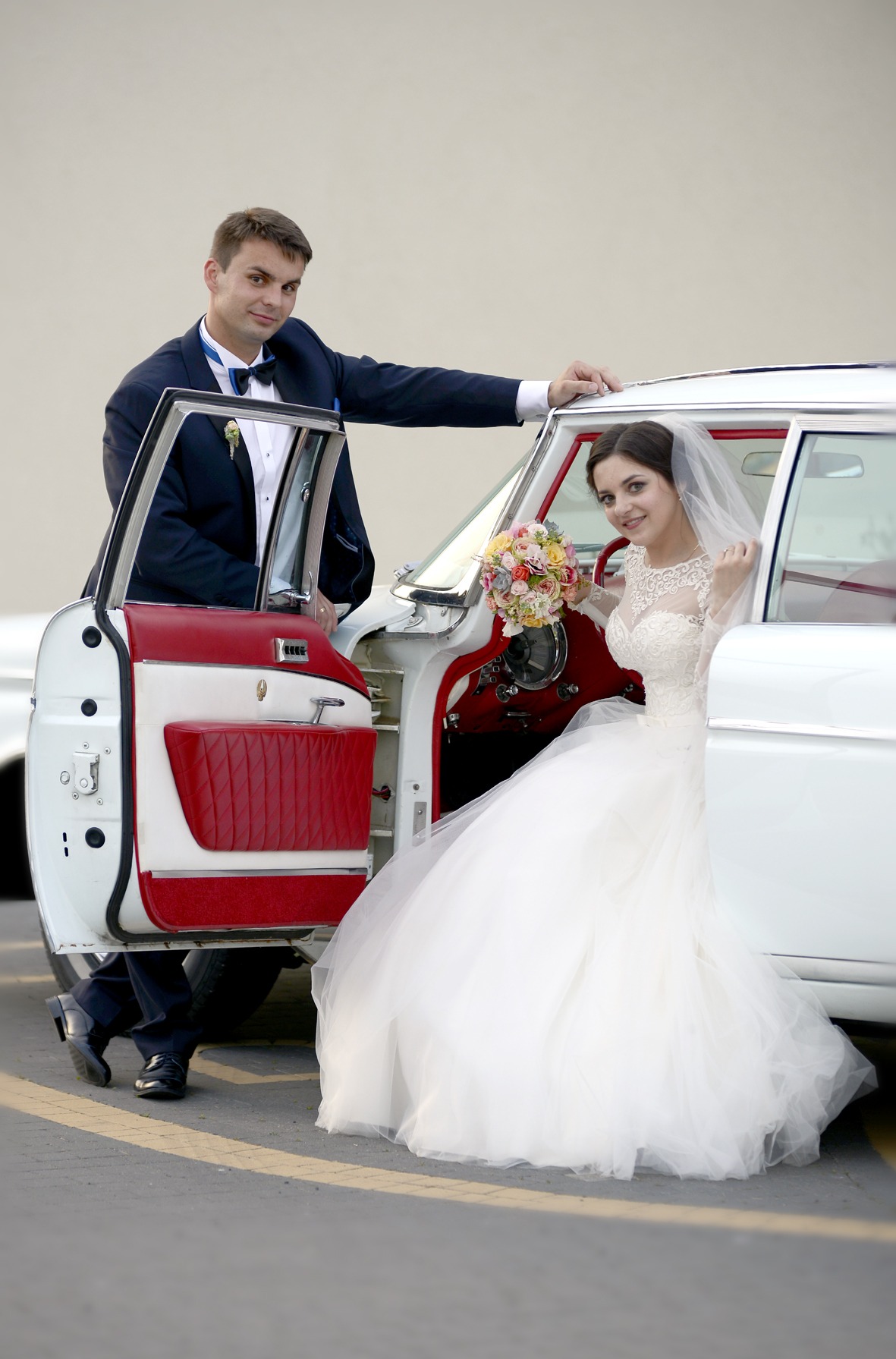 Uśmiechnięta para młoda pozuje przy białym, klasycznym samochodzie z czerwonym wnętrzem. Panna młoda w sukni z bukietem, pan młody w granatowym garniturze.