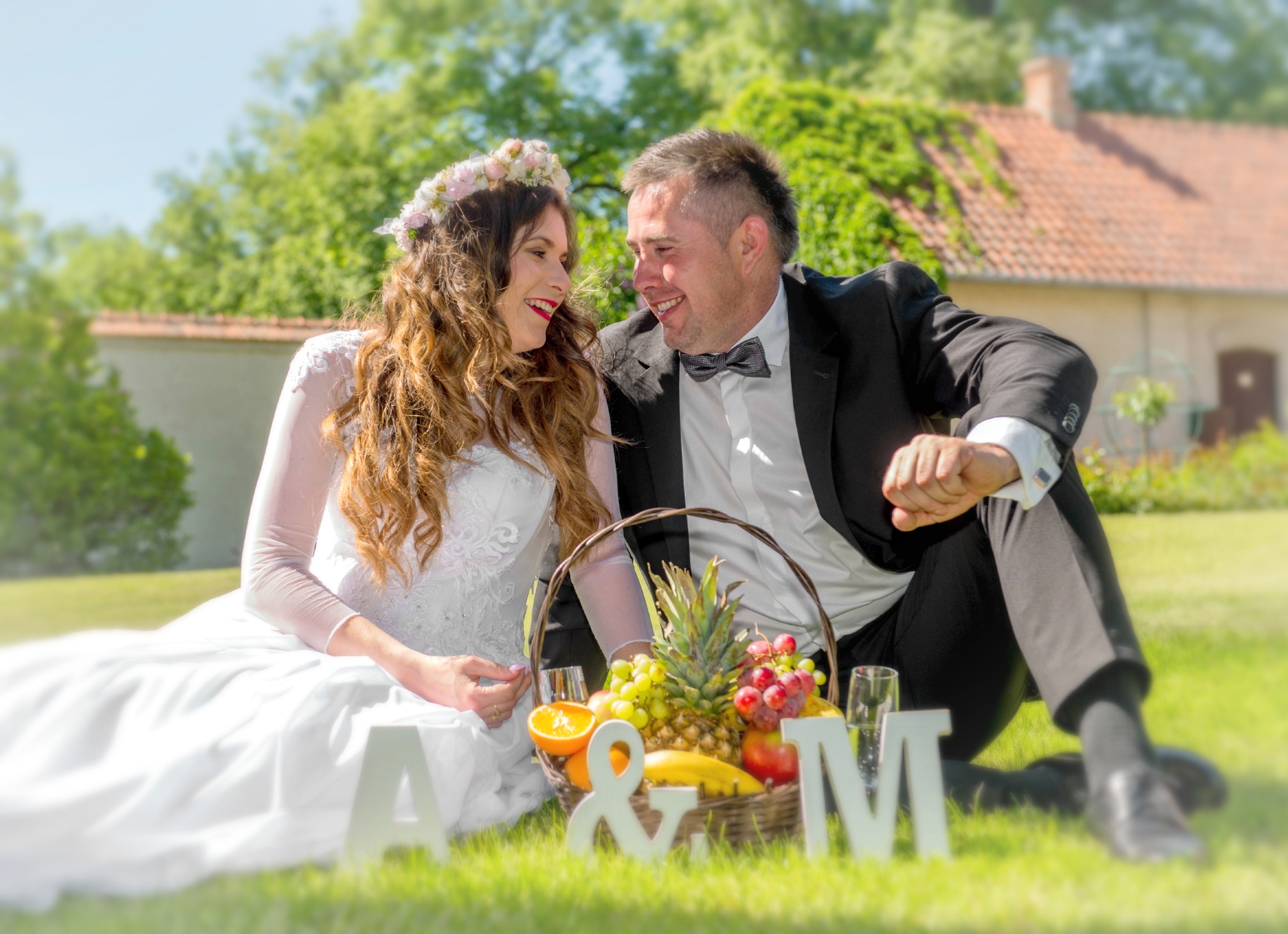 Uśmiechnięta para młoda siedzi na trawie obok kosza z owocami i liter A&M w słoneczny dzień. Panna młoda ma wianek na głowie, a pan młody muchę.